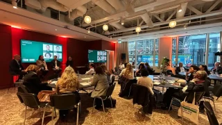 Workshop participants are seated at various tables, facing the front of the room where the speakers/panelists are seated; behind the speakers are two screens in green which feature their names.